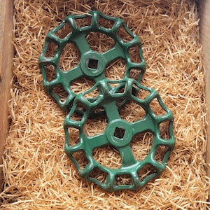 Puede incluir: Dos manijas de válvula de hierro fundido verde con un diseño circular y un centro cuadrado. Las manijas están colocadas en una caja de madera llena de paja. Las manijas tienen una estética vintage e industrial.