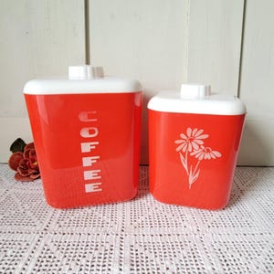 May include: Two rectangular storage canisters with rounded corners, featuring a bright red body and a white lid. The larger canister has the word COFFEE printed vertically in white letters, while the smaller canister displays a white floral graphic.