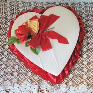 May include: A heart-shaped candy box with a white lid, decorated with a red satin bow and a red fabric rose. A heart-shaped tag attached to the rose reads Russell Stover Candies, Valentine, Net Wt 20 oz (1 lb 4 oz). The box has red ruffled trim.