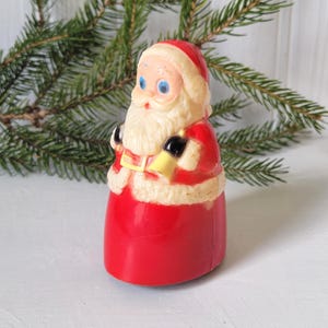 May include: A molded plastic Santa Claus figurine featuring a bright red suit with white trim, a white beard, and blue eyes. The figure holds a small yellow bell in one hand and wears a red hat with a white brim, standing in a vertical orientation.