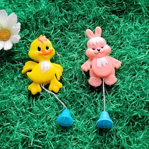May include: Two plastic Easter-themed toys: a yellow duckling and a pink bunny. Each figure has a string attached to a blue bell. The toys are set against a green, grassy background, with a white daisy in the upper left corner.