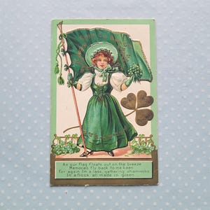 Puede incluir: Postal vintage del Día de San Patricio con una mujer con un vestido y sombrero verdes sosteniendo una bandera y tréboles. La postal tiene una combinación de colores verde y blanco con el texto "Dear Irish Memories."