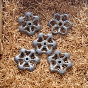 Puede incluir: Cinco manijas de grifo vintage de color plateado con un diseño de flor de seis pétalos. Las manijas están dispuestas en una caja de madera llena de paja. Las manijas tienen un centro circular y probablemente son de metal.