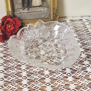 Puede incluir: Un cuenco de vidrio transparente con un diseño de hojas y bayas. El cuenco tiene forma ovalada con una superficie texturizada. El cuenco está sobre un mantel de encaje blanco. Una flor roja y una foto enmarcada están en el fondo.