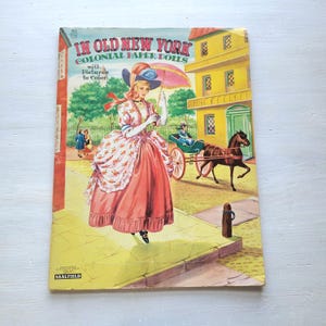 May include: Vintage "In Old New York Colonial Paper Dolls" book cover. The cover features a woman in a colonial dress holding a parasol, with a horse-drawn carriage and a yellow building in the background. The cover is slightly worn.