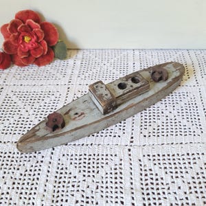 May include: A weathered, light blue wooden toy boat with a distressed finish. The boat features a central rectangular cabin structure with two circular openings and two small, dark brown circular wooden components attached to the deck.