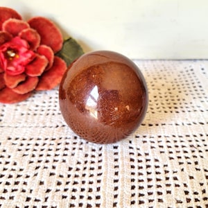 May include: A smooth, polished, spherical object with a deep reddish-brown, marbled appearance. The surface reflects light, showing subtle variations in color and texture across its rounded form, positioned near a large red flower.