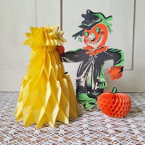 May include: A vintage-style paper scarecrow cutout with a bright orange head, black hat, and green clothing, accompanied by a yellow honeycomb paper decoration and a small orange honeycomb paper pumpkin, all featuring a classic autumn aesthetic.