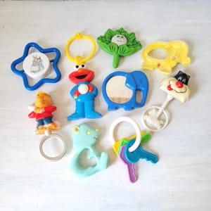 Peut inclure: Ensemble de jouets pour bébés vintage colorés. On y trouve un hochet bleu en forme d'étoile avec une image de Pierre Lapin, une figurine d'Elmo, un miroir éléphant bleu et un hochet Sylvestre le Chat. Autres jouets : une feuille verte et un porte-clés.