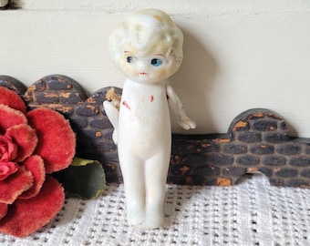 Könnte beinhalten: Eine kleine, cremeweiße Porzellanpuppe mit modelliertem blondem Haar, blau bemalten Augen und roten Lippen. Die Puppe hat eine glatte, blasse Oberfläche mit kleinen roten Farbmarkierungen auf Brust und Schulter sowie bewegliche, abstehende Arme.