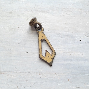 Puede incluir: Tirador de cajón antiguo de latón con un diseño geométrico decorativo. El tirador tiene forma de diamante con un recorte y una pequeña parte superior texturizada. El metal tiene un aspecto desgastado y envejecido.