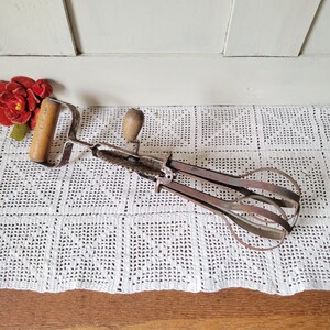 May include: Vintage hand-crank egg beater with wooden handles and a rusty metal frame. The beater is resting on a white crocheted tablecloth, with a red flower in the background. A classic kitchen tool.