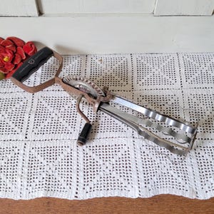 May include: Vintage hand-crank egg beater with a black handle and a metal whisk. The beater has a rusty, aged appearance, suggesting it's an antique kitchen tool. It rests on a white, crocheted tablecloth, with a red flower arrangement in the background.