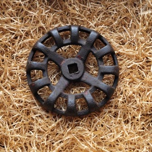 Puede incluir: Una rueda de válvula circular de metal oscuro, con un orificio central cuadrado y aberturas rectangulares. La rueda tiene un aspecto envejecido y desgastado, con rastros de óxido rojo. Descansa sobre virutas de madera marrón claro.