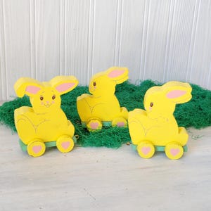 May include: Three yellow wooden bunny figurines with pink accents, each on a green base with pink heart-shaped wheels. The bunnies have large ears and are set against a backdrop of green moss and a white wooden surface.