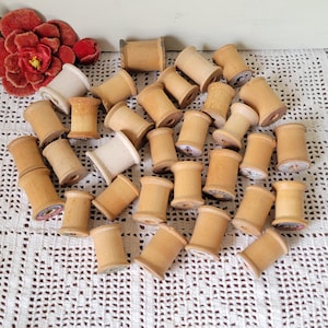 Pode incluir: Uma coleção de numerosas bobinas de madeira vazias em vários tons de castanho claro e bege, espalhadas sobre uma superfície de crochet branca. Uma grande flor de tecido vermelho escuro está posicionada no canto superior esquerdo.