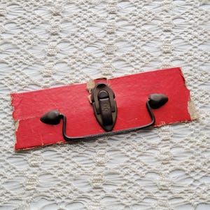 Puede incluir: Un tirador de metal oscuro con una placa de montaje central que presenta un mecanismo de cierre vertical. El herraje está sujeto a un trozo rectangular de cartón rojo con bordes deshilachados, mostrando los puntos de montaje y el asa curva.