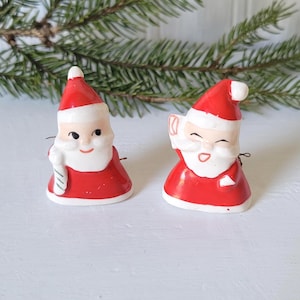 May include: Two vintage Santa Claus figurines. Each figurine is wearing a red suit and hat with white trim and a white beard. One holds a candy cane, the other a gift. The figurines are on a white surface with evergreen branches in the background.