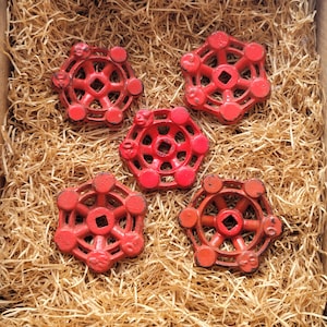 Puede incluir: Cinco manijas de válvula rojas de estilo vintage con un diseño hexagonal, dispuestas sobre un lecho de paja. Las manijas tienen un centro circular y están rodeadas de pequeños acentos circulares. La estética general es rústica e industrial.