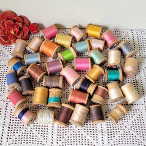 Può includere: Una collezione di rocchetti di filo da cucito assortiti in vari colori, tra cui rosa, giallo, blu, marrone e bianco, ciascuno avvolto su un piccolo cilindro di legno. Un grande fiore in tessuto rosso scuro è posizionato nell'angolo in alto a sinistra.