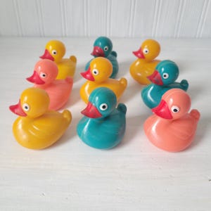 May include: A collection of colorful rubber duck toys in various shades of yellow, teal, and coral. Each duck has a red beak and white eyes. The ducks are arranged on a white surface.