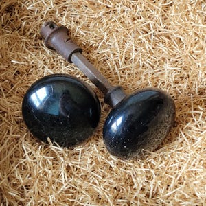 May include: Two antique black door knobs with a connecting rod. The knobs are round and oval-shaped, with a glossy finish. The connecting rod is made of metal and has a dark, aged appearance. The knobs are set against a background of straw.