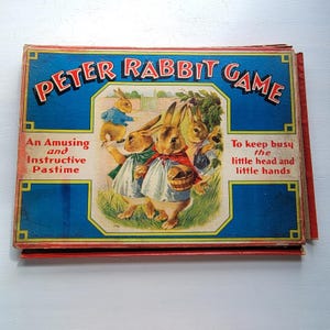 Puede incluir: Caja de juego vintage de Peter Rabbit con fondo azul y letras rojas. La caja presenta ilustraciones de conejos vestidos, con el texto "An Amusing and Instructive Pastime" y "To keep busy the little head and little hands."