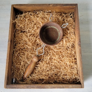 May include: A small, round, brown sieve with a wooden handle and metal frame rests on a bed of straw inside a wooden box. The sieve is centered in the box, with the handle extending towards the bottom left corner. The box is made of aged wood.