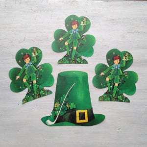 Könnte beinhalten: St. Patrick's Day Dekorationen. Ein grüner Leprechaun-Hut mit goldener Schnalle und drei Leprechaun-Figuren, die auf Kleeblättern stehen. Die Figuren tragen grüne Anzüge und Fliegen. Die Dekorationen befinden sich auf einer hellgrauen Oberfläche.