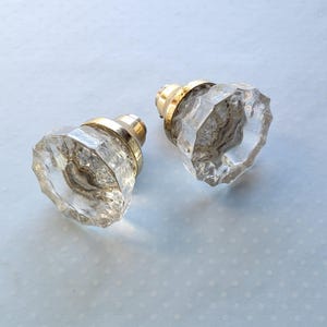 May include: Two clear, faceted glass door knobs with gold-colored metal bases. The knobs have a vintage aesthetic and are likely for interior doors or cabinets. The glass is transparent, allowing light to pass through.