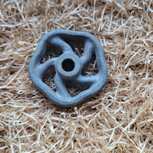 Puede incluir: Una pequeña rueda de metal gris desgastado con un agujero central y seis radios. La rueda tiene un aspecto rústico e industrial y está colocada sobre un fondo de paja.