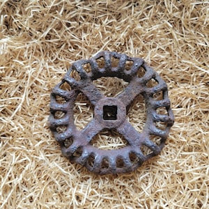 Puede incluir: Un manillar de válvula circular de metal oxidado con un centro cuadrado y una serie de cortes calados alrededor del perímetro. El manillar está sobre un fondo de material de embalaje triturado de color marrón claro.