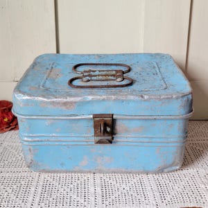 May include: A rectangular light blue metal storage box with a distressed finish showing silver-toned metal underneath. The box features a rusted metal handle on the lid and a rusted metal latch on the front for securing the lid in a closed position.