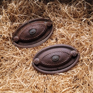 Puede incluir: Dos tiradores de cajón ovalados de estilo antiguo con un acabado marrón óxido desgastado. Cada tirador presenta un medallón central decorativo y pequeños pomos en los extremos. Los tiradores se exhiben sobre un lecho de paja.