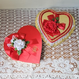 May include: Two heart-shaped boxes. One is red with a white flower, red ribbon, and a heart-shaped tag reading Valentine Greetings. The other is gold with a red ribbon, a red flower, and a white lace trim border around the edge of the heart.