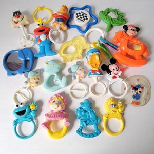 Op de afbeelding: Een verzameling vintage baby rammelaars in verschillende vormen en kleuren. De rammelaars bevatten stripfiguren zoals Mickey Mouse, Donald Duck en Cookie Monster. Andere vormen zijn een ster, een schommelpaard en een bloem.