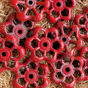 Puede incluir: Un conjunto de manijas de válvula vintage rojas en forma de flor con un acabado envejecido. Las manijas están dispuestas sobre un lecho de paja, mostrando su apariencia envejecida y su diseño único. Estas piezas decorativas añaden un encanto rústico.