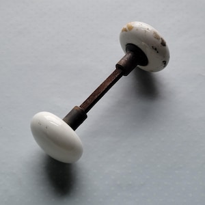 May include: An antique door knob with white ceramic oval handles and a rusty brown metal rod. The knobs have a glossy finish with brown and yellow speckles. A classic hardware piece.