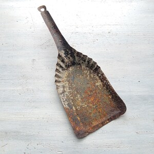 May include: A vintage metal dustpan, showing signs of rust and wear, with a long handle and a small hanging hole. The dustpan has a decorative, scalloped edge. The colours are a mix of brown, grey, and orange.