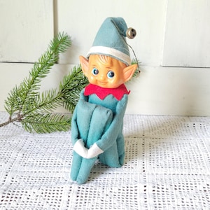 Op de afbeelding: Een pluche elfpop met een blauwgroen lichaam, een bijpassende puntmuts met een klein metalen belletje en een rode kraag. De pop heeft een gegoten kunststof gezicht met blauwe ogen, puntige oren en witte boorden aan de mouwen en broekspijpen.