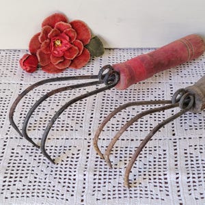 May include: Two hand cultivators with curved metal tines and wooden handles. One handle is painted red, while the other is natural wood. A large, deep red fabric flower with green leaves is positioned near the red-handled tool.