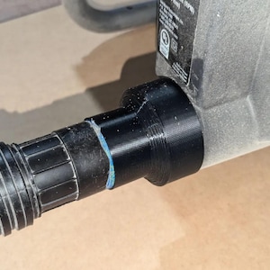 May include: A close-up shot of a black vacuum hose connected to a grey power tool. The hose is ribbed and attached to the tool with a black adapter. The tool has a label with text and a power cord.