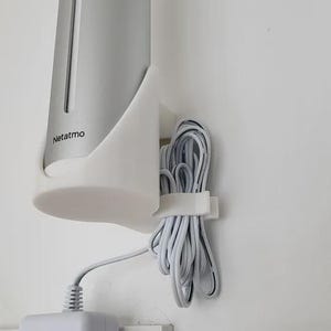 May include: A light grey Netatmo weather station, mounted on a white wall. The device is held in a white bracket, with a white power cord and adapter plugged into a wall socket. The Netatmo logo is visible on the device.