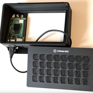 May include: A black Stream Deck control panel with 24 square buttons, next to a black enclosure with a Raspberry Pi circuit board. The Stream Deck has the text "STREAM DECK" printed on it. A black USB cable connects the two components.