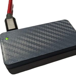 May include: A dark grey rectangular device with a carbon fibre pattern, connected to a red cable with a silver connector. A small green light is illuminated on the device, indicating it is powered on.