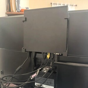 May include: A black device attached to the rear of a flat-screen television, with multiple cables and connectors connected. The device seems to be a mounting bracket or housing for electronic components. The TV is black.
