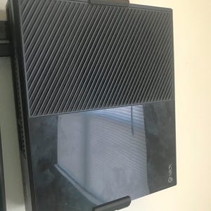 May include: A black Xbox One console mounted on a wall with a black bracket. The console has a textured top with diagonal lines and a glossy bottom. The Xbox logo is visible on the lower right corner.