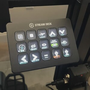 May include: Black Stream Deck with illuminated buttons displaying icons for flight controls, navigation, and radio functions. The Stream Deck is mounted on a black frame, with a cable connected at the bottom.