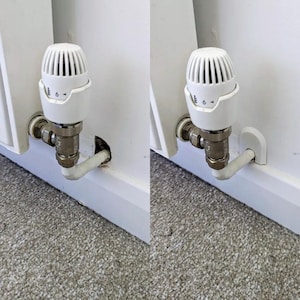May include: Two white radiator valves, set to 6, are mounted on a white wall. They connect to a metal pipe and a white pipe. The background features a white wall and a grey carpet.