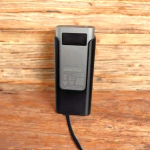 May include: A silver and black electronic device, possibly a charger or a portable media player, is shown in a black holder. The device has a small black screen and a cable extending from the bottom. The background is a wooden surface.